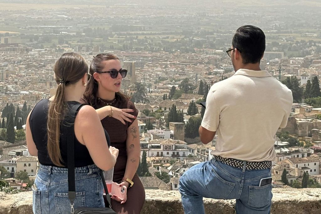 한 명은 선글라스를 쓰고 다른 한 명은 배낭을 메고 도시 풍경 앞에서 이야기를 나누는 두 명의 여성. 도시를 배경으로 한 옥상 풍경.