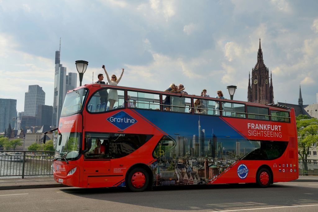 도시 건물과 가로등 기둥으로 둘러싸인 꼭대기 갑판에 사람들이 있는 밝은 빨간색 2층 관광 버스.