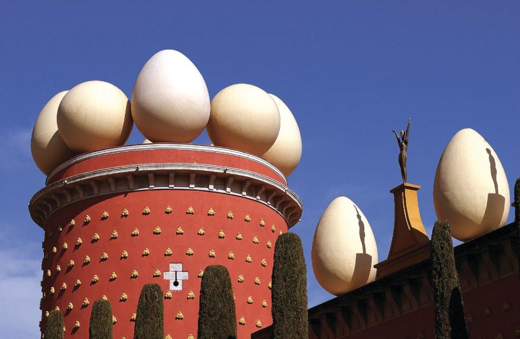 붉은 건물 위에 커다란 계란 조각이 있고, 맑고 푸른 하늘 아래 옥상 모퉁이에 서 있는 사람의 동상이 있습니다.