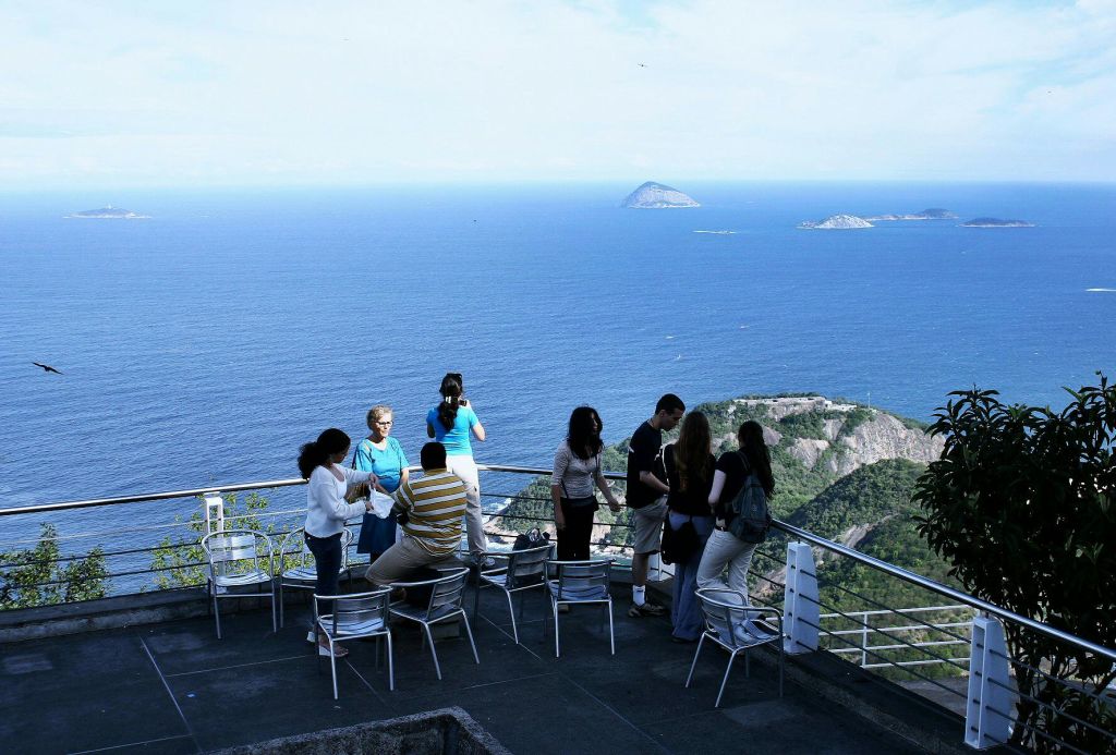 넓은 바다가 내려다보이는 테라스에 한 무리의 사람들이 서 있으며, 멀리 작은 섬들이 몇 개 있습니다.