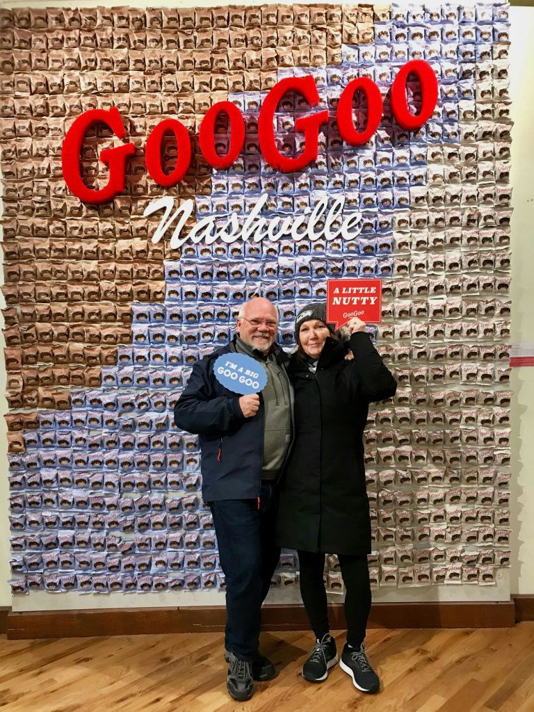 포장된 Goo Goo Clusters가 전시된 벽 앞에서 두 사람이 포즈를 취하고 있으며, 위에는 "Goo Goo Nashville" 표지판이 있습니다.