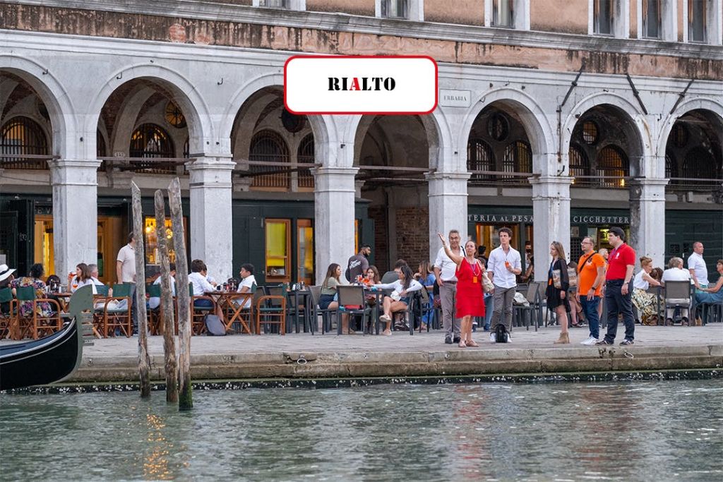 아치형 창문과 운하 옆의 '리알토' 라고 쓰인 표지판이 있는 오래된 건물 근처에 사람들이 서서 앉아 있습니다.