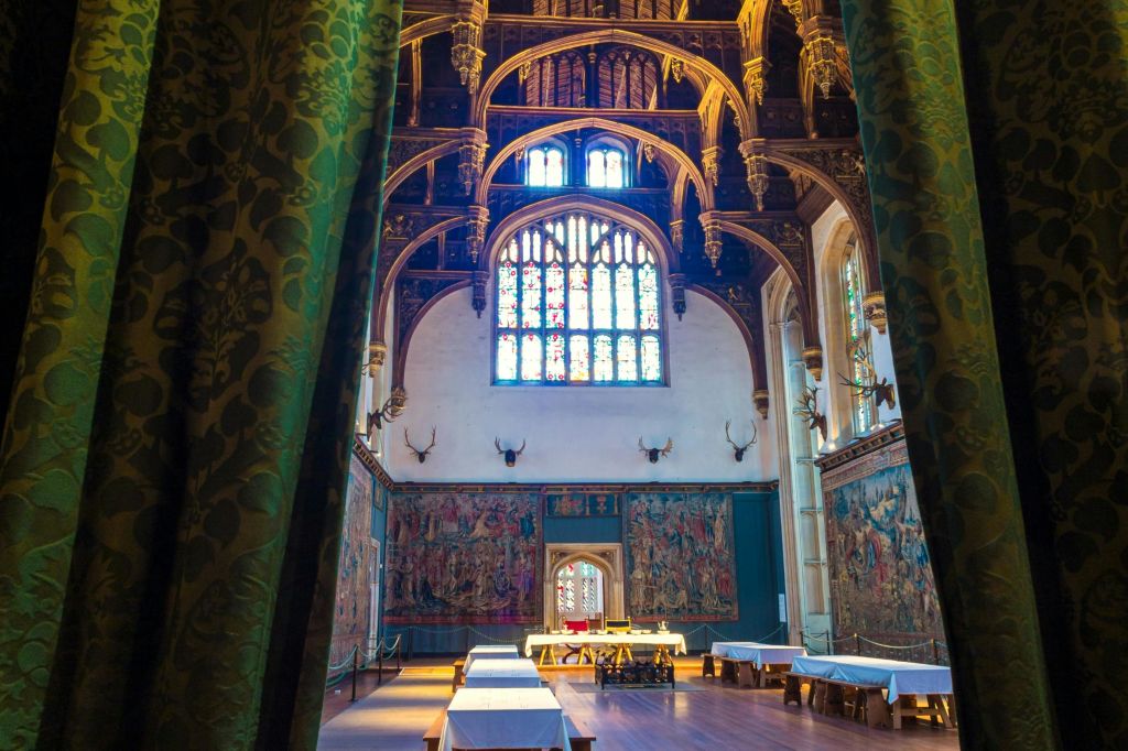 아치형 천장, 스테인드글라스 창문, 뿔 장식, 벽에 태피스트리, 천으로 덮인 긴 테이블이 있는 화려한 홀.