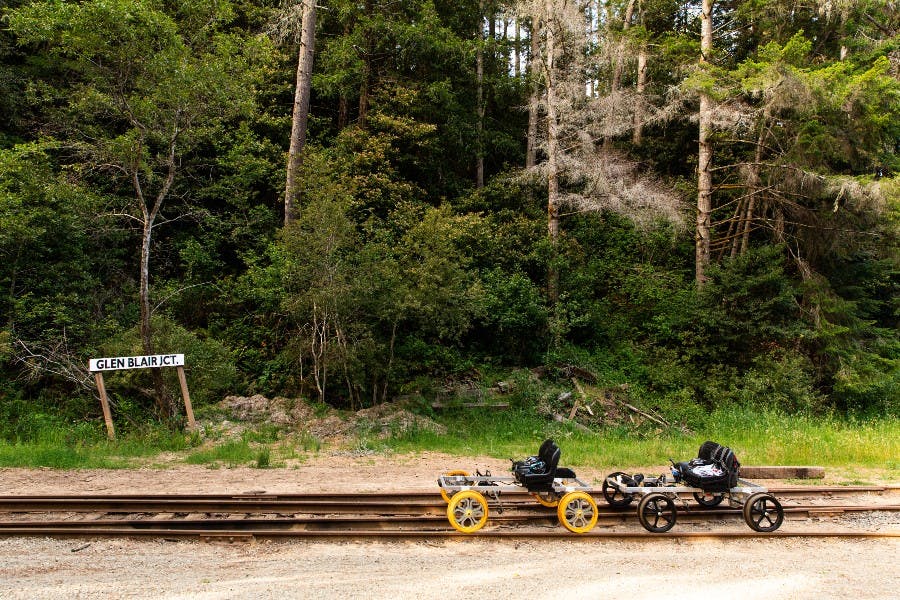 좌석과 페달을 갖춘 두 개의 철도 카트가 울창한 숲 앞의 기차 트랙에 주차되어 있습니다. 표지판에는 "R.R. JCT" 라고 쓰여 있습니다.