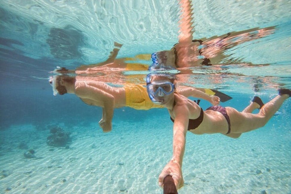 두 사람이 수중에서 스노클링을 하고 있습니다. 한 명은 셀카봉을 사용하여 비키니를 입고, 다른 한 명은 노란색 수영복을 입고 있습니다. 맑은 물과 모래 해저가 보입니다.