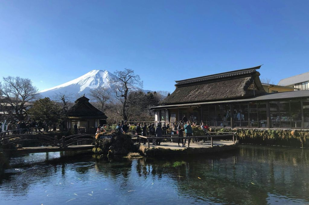 반사되는 고요한 연못, 초가 지붕이 있는 목조 건물, 맑은 하늘 아래 후지산을 배경으로.