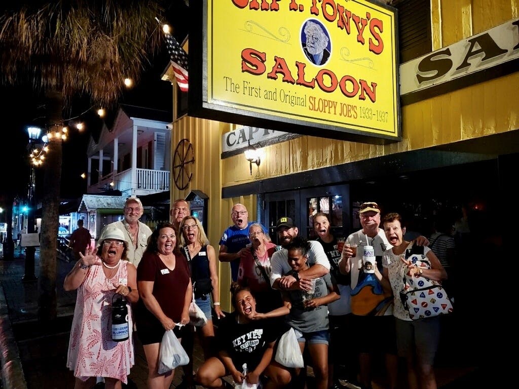 한 무리의 사람들이 건물 앞에서 "Capt. Tony's Saloon" 이라고 적힌 간판을 들고 행복한 포즈를 취하고 있습니다.