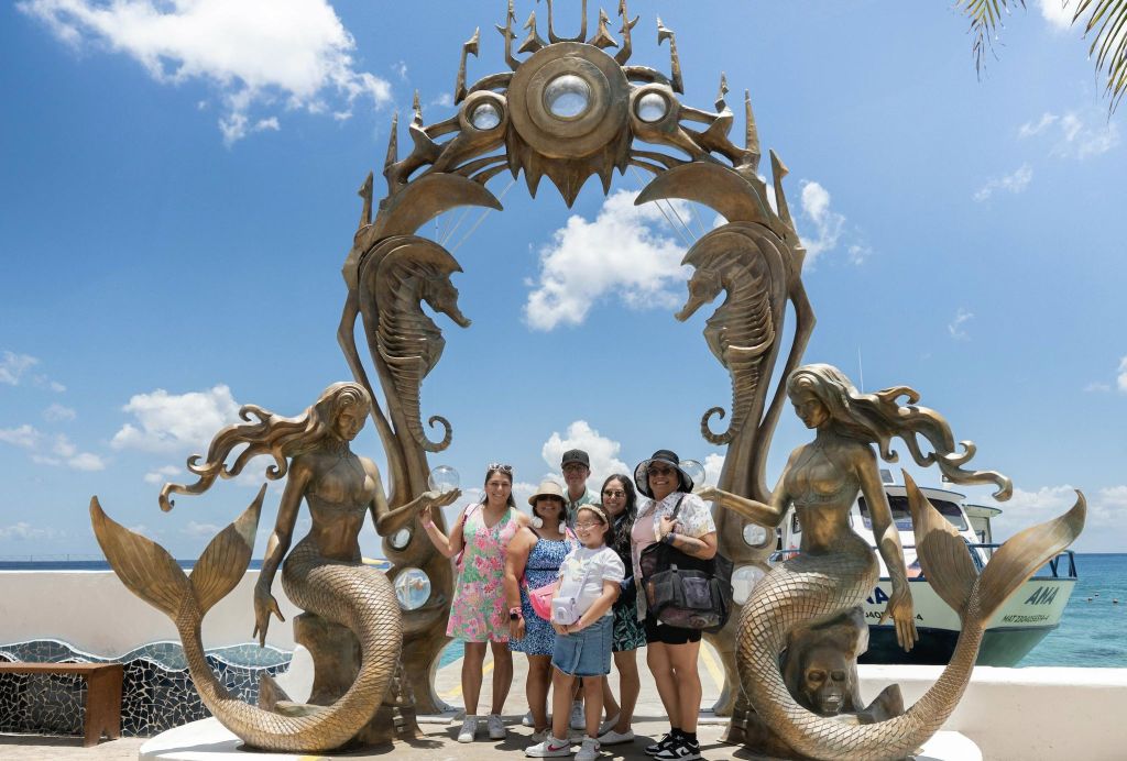 인어와 해마로 장식된 크고 화려한 아치 아래에는 푸른 하늘과 구름을 배경으로 여섯 명의 사람들이 서 있습니다.