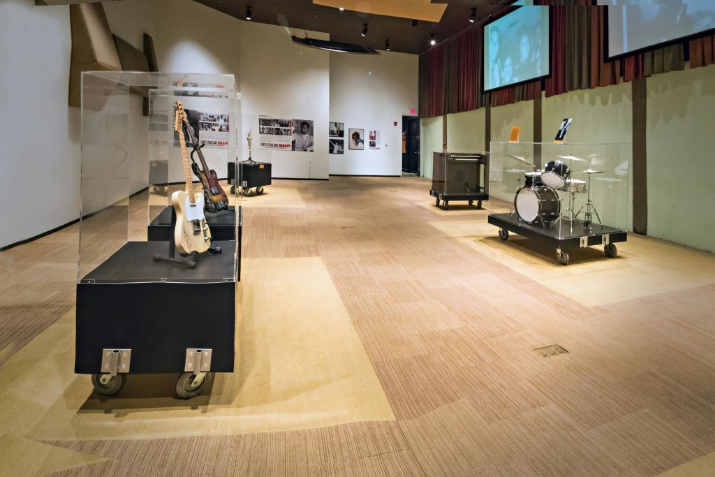 일렉트릭 기타, 바퀴 달린 플랫폼의 드럼 키트, 부드러운 조명 아래 벽에 걸린 빈티지 음악 포스터가 있는 박물관 전시.