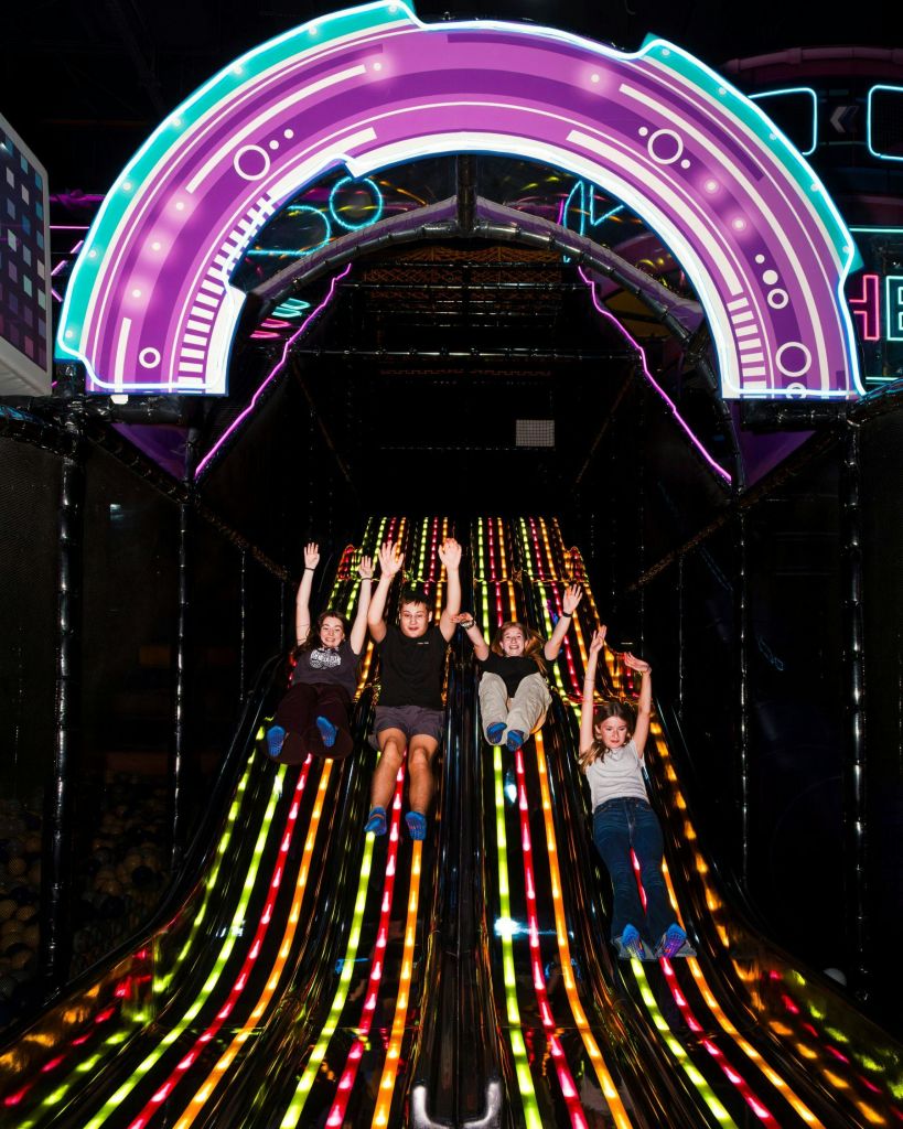네 명의 어린이가 다채롭고 빛나는 슬라이드를 아래로 미끄러지듯 내리면서 양손을 들어 올립니다. 배경에는 네온 조명과 아케이드 장식이 보입니다.