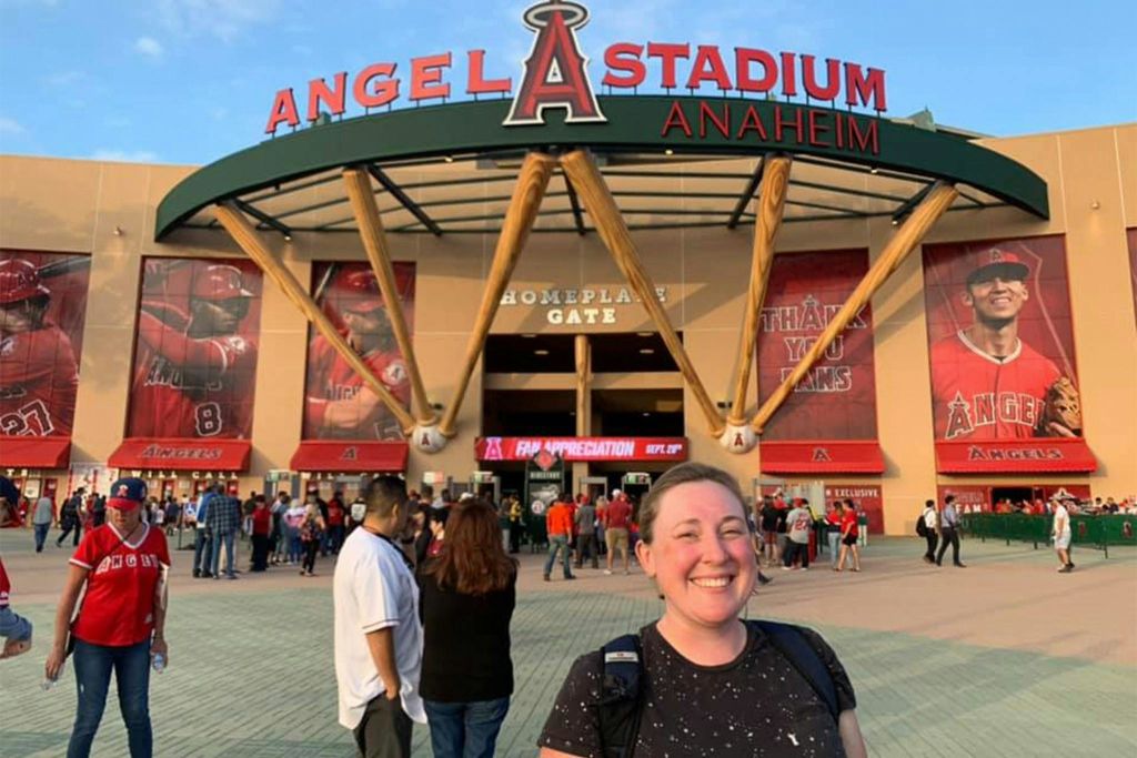 애너하임의 엔젤 스타디움 앞에서 미소를 짓고 있는 한 사람이 간판이 보이고 사람들이 입장하고 있습니다.