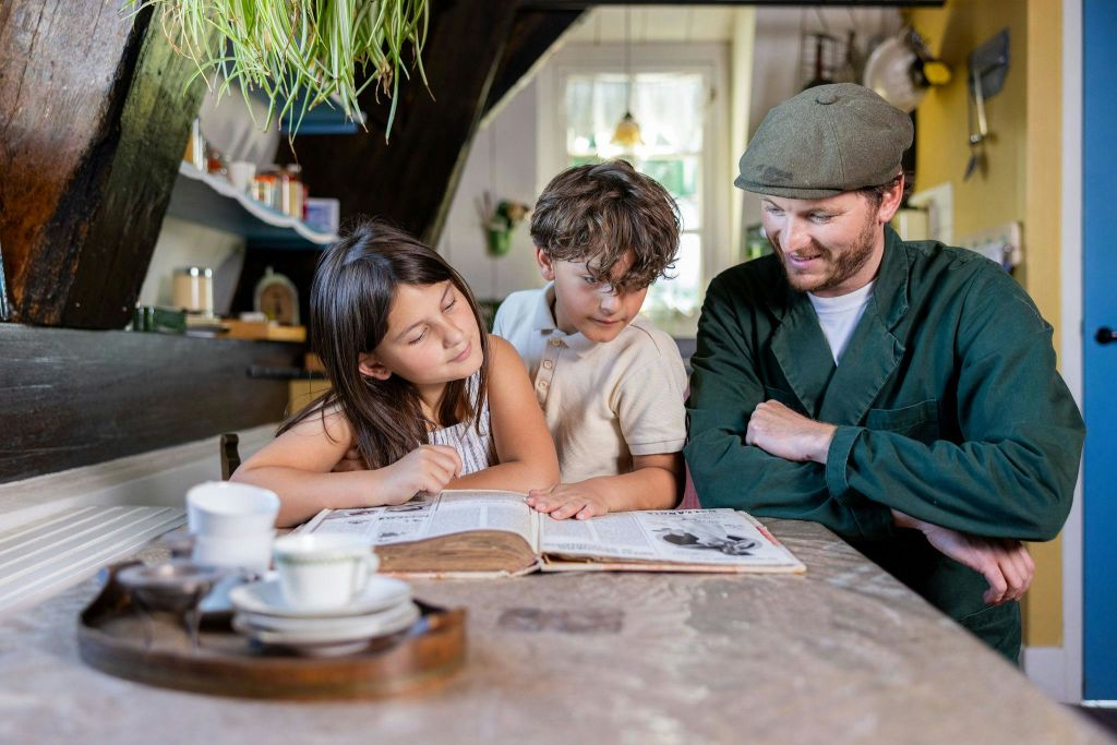 한 남자와 두 자녀가 테이블에 앉아 아늑한 주방에서 함께 열린 책을 보고 있습니다.