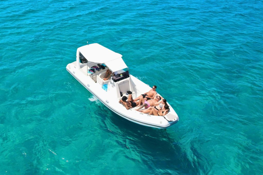 햇살이 내리쬐는 하늘 아래 맑은 청록색 바다에서 하얀 보트를 타고 휴식을 취하고 있는 한 무리의 사람들.