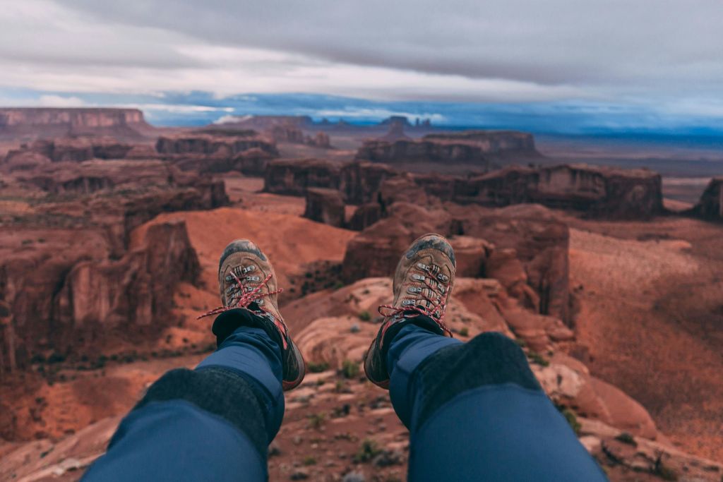 푸른 바지를 입은 하이킹 부츠와 다리가 광활한 사막 협곡 풍경에 매달려 흐린 하늘 아래 암석이 형성되어 있습니다.