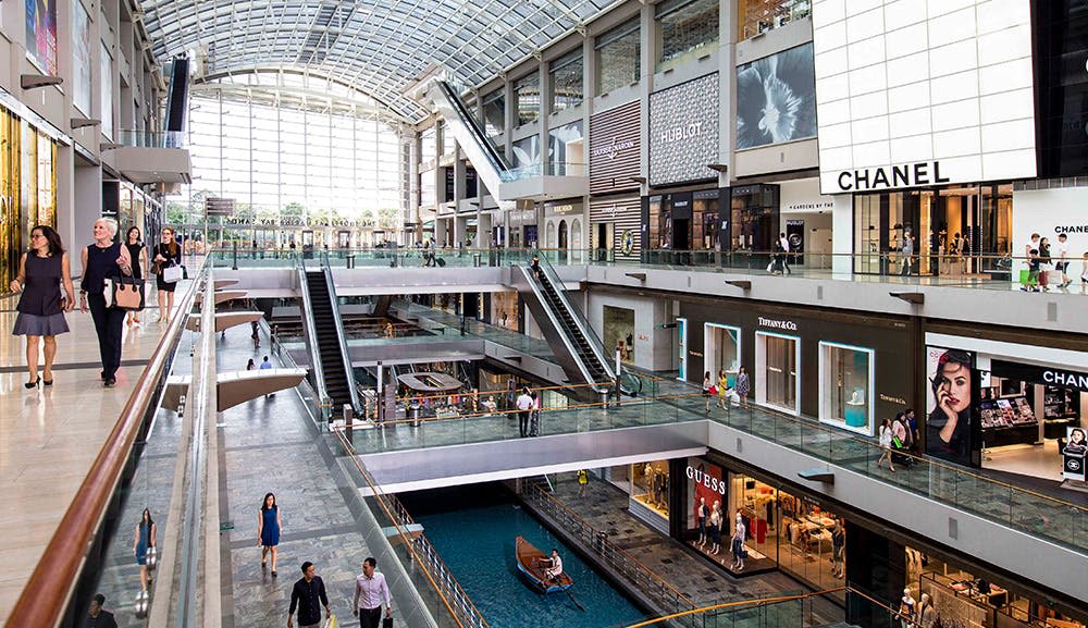 위블로, 샤넬, 게스와 같은 매장이 있는 3층 쇼핑몰. 쇼핑객들이 걷고 있고 보트가 실내 운하에 있습니다.