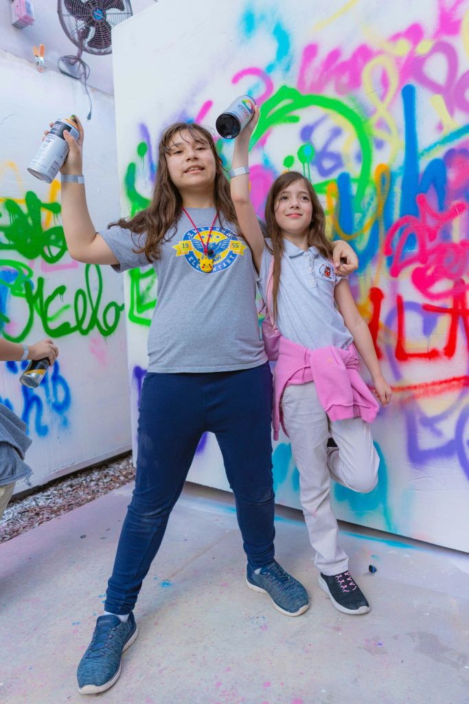 스프레이 페인트 캔을 들고 있는 두 명의 소녀가 그래피티로 덮인 벽 앞에 서서 웃으며 팔을 들어 올린 채 포즈를 취하고 있습니다.