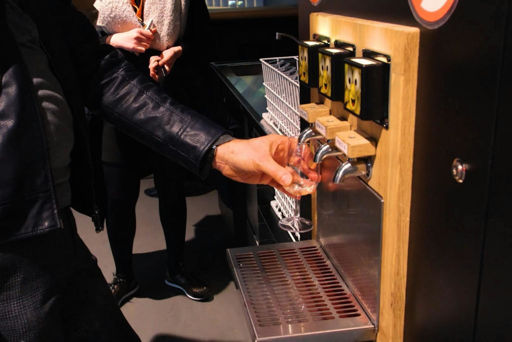 가죽 재킷을 입은 사람이 공공 장소에서 손잡이에 만화 얼굴이 있는 맥주 탭에서 잔을 채우고 있습니다.