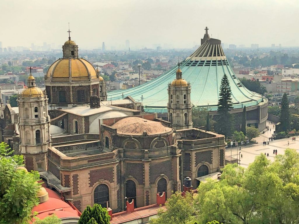 도시 경관을 배경으로 경사진 지붕이 있는 돔과 현대적인 바실리카가 있는 역사적인 교회입니다. 전경의 나무와 안개로 희미해진 스카이라인.