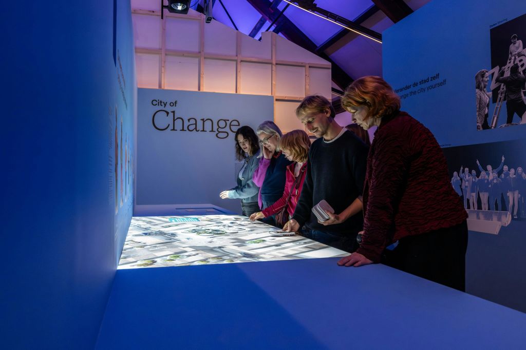 벽에 "변화의 도시" 가 있는 현대적인 전시 공간에서 조명이 켜진 대화형 디스플레이 테이블을 살펴보는 사람들.