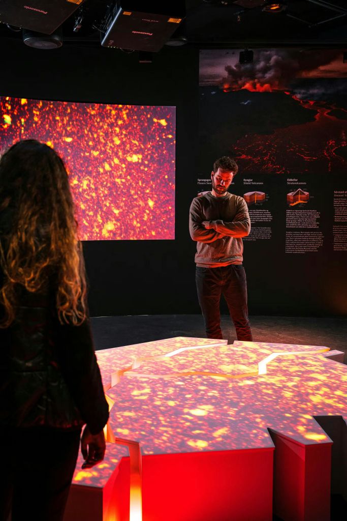 어두운 방에 있는 두 사람이 불타는 패턴과 정보 패널을 특징으로 하는 빛나는 대화형 전시물을 관찰합니다.