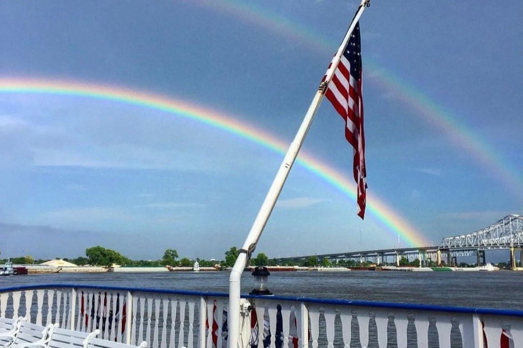 다리를 배경으로 강을 건너는 이중 무지개. 전경의 보트 갑판에 미국 국기가 있습니다.