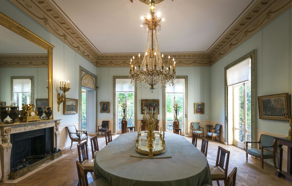 커다란 샹들리에가 있는 우아한 다이닝 룸, 장식용 센터피스가 있는 타원형 테이블, 벽에 놓인 의자와 그림으로 둘러싸여 있습니다.