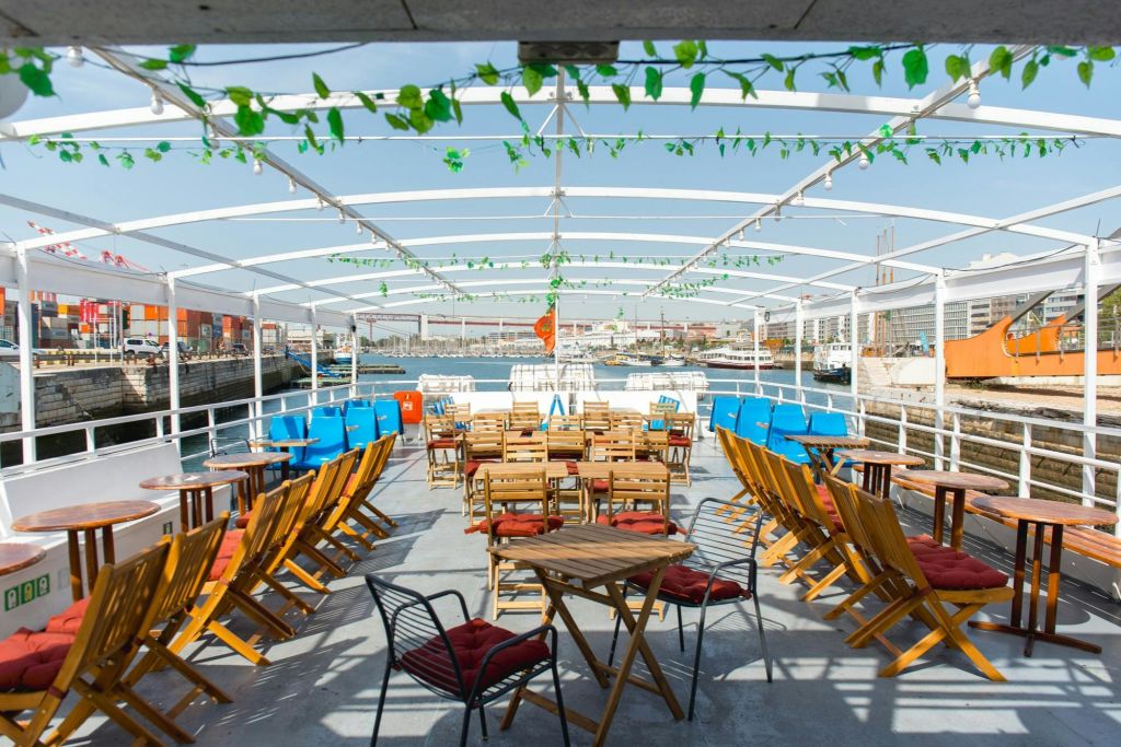 항구가 내려다보이는 녹색 덩굴로 장식된 나무 의자와 테이블이 있는 보트 데크의 개방형 좌석 공간.