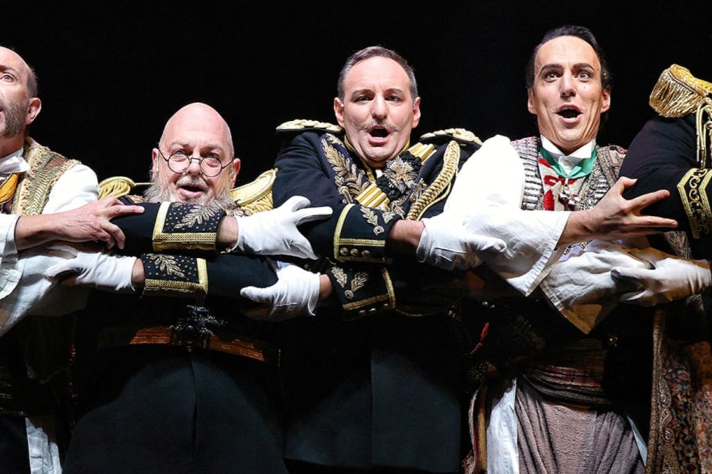 무대에서 공연하는 정교한 의상을 입은 세 남자가 노래하거나 일제히 연기를 제안하는 표정을 짓고 있습니다.