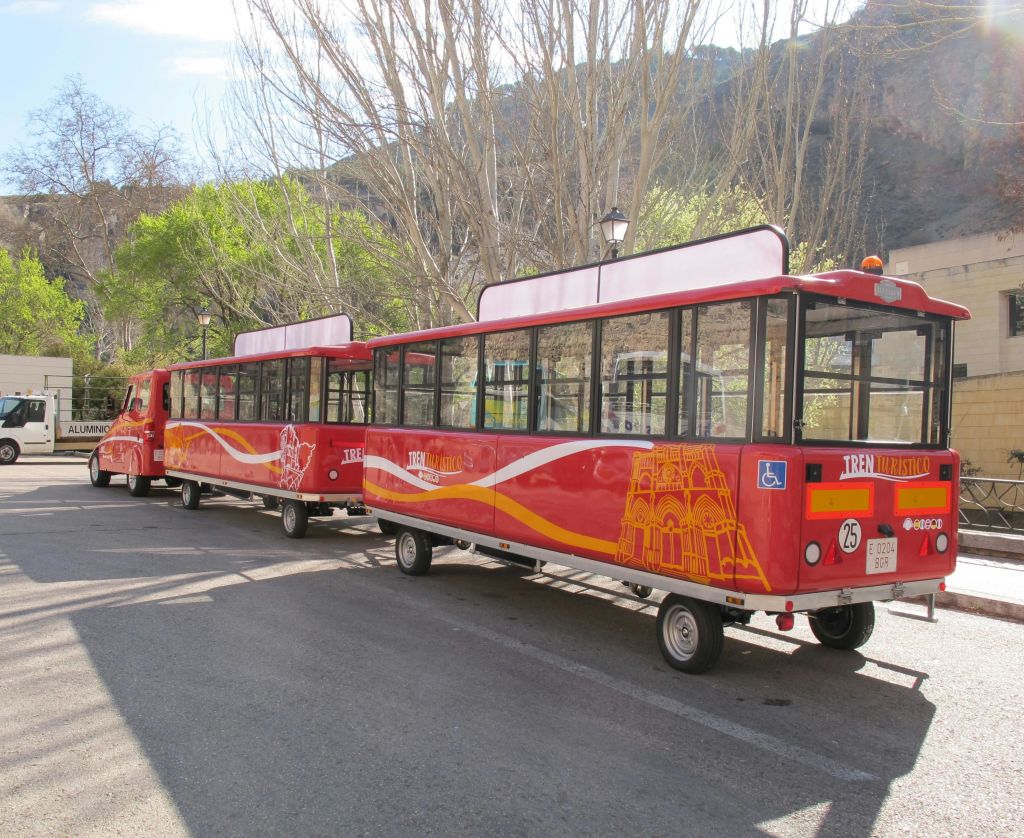 맨발의 나무와 맑은 하늘 아래 녹지 근처의 거리에 두 대의 마차가 주차된 빨간색 관광 열차입니다.