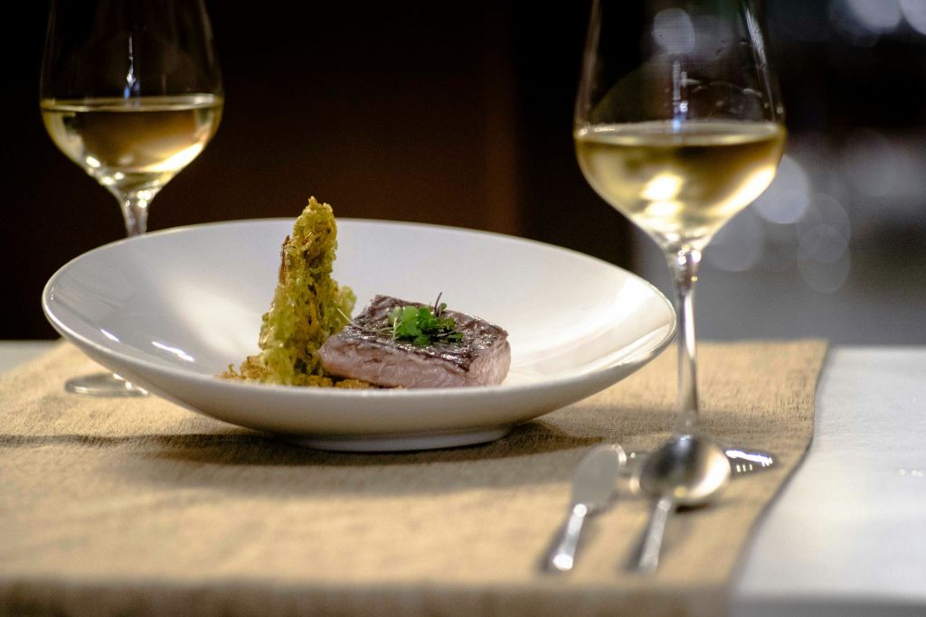 허브, 화이트 와인 2잔, 베이지색 식탁보 위에 식기류가 장식된 도금 고기 요리.