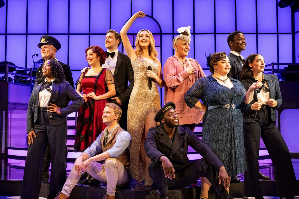 다양한 다채로운 의상을 입고 밝은 조명 아래에서 미소를 짓고 포즈를 취하는 무대의 사람들. 중앙에 있는 한 여성이 마이크를 들고 있습니다.