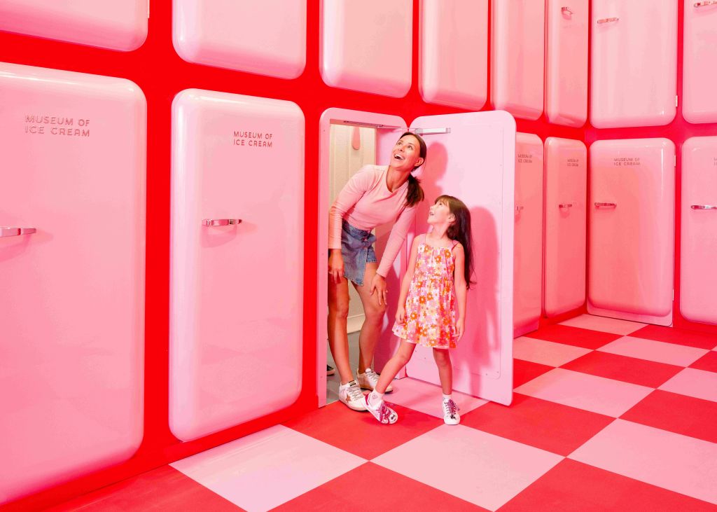 아이스크림을 테마로 한 인테리어가 돋보이는 분홍색 객실의 두 사람이 탁 트인 분홍색 냉장고, 빨간색과 분홍색 체크무늬 바닥 옆에 서 있습니다.