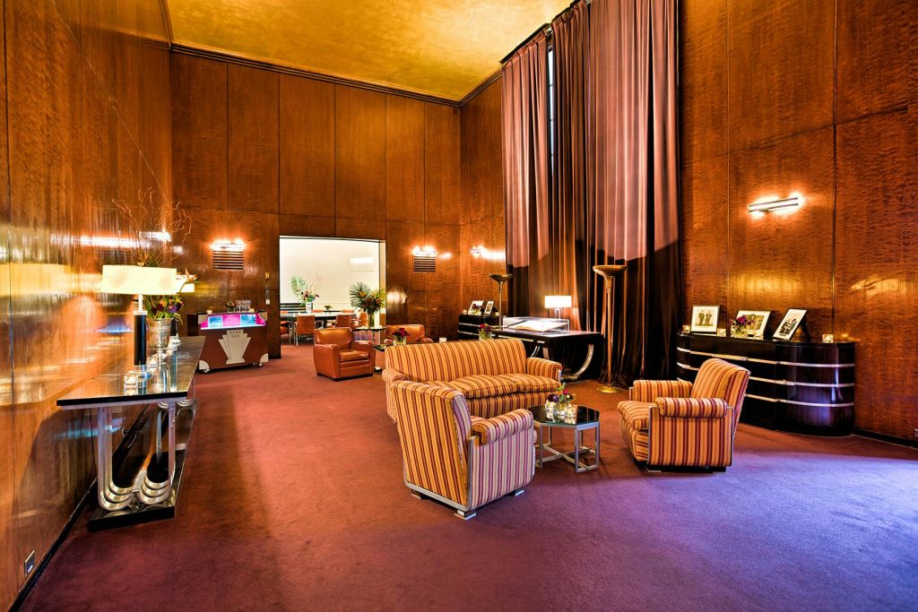 줄무늬 소파, 벨벳 커튼, 나무 패널, 주변 조명이 있는 고급스러운 라운지입니다. 배경에 인접한 방이 보입니다.