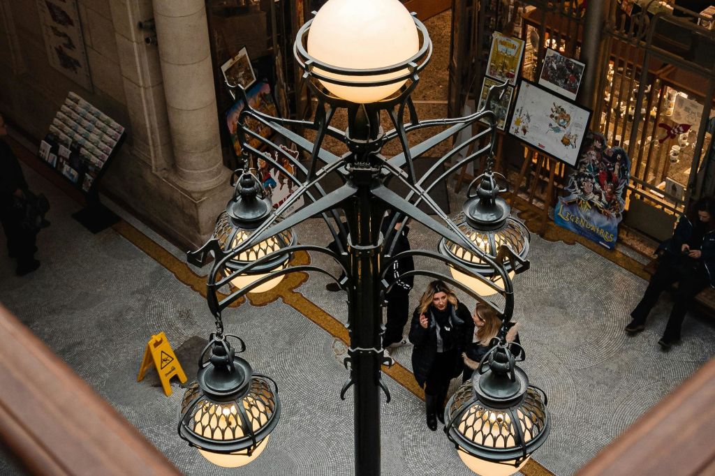 건물 내부의 장식용 가로등 근처에 사람들이 서 있습니다. 예술품이 근처에 전시되어 있으며, 바닥에 주의 표지판이 보입니다.