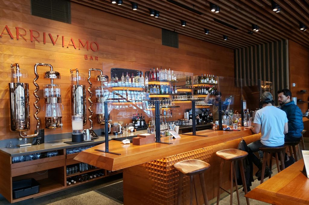 높은 스툴, 선반에 놓인 다양한 주류 병, 플렉시글라스 칸막이 뒤에 있는 커피 장비가 있는 나무 바.