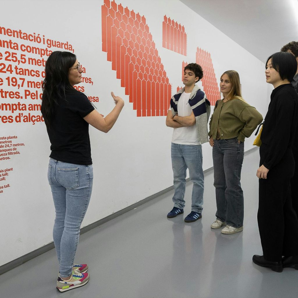 한 여성이 세 명의 여성과 한 명의 남성으로 구성된 그룹과 이야기하는 동안 몸짓을 하고 있으며, 흰색 벽 뒤에 빨간색 텍스트와 그래픽이 있습니다.