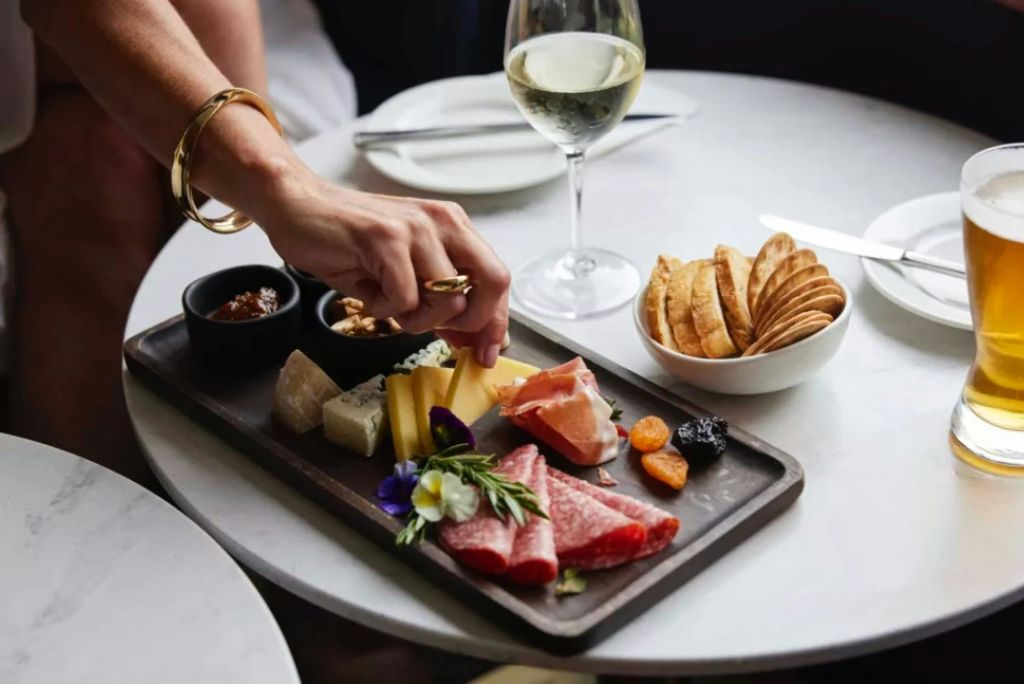 고기, 치즈, 과일이 있는 샤퀴트리 보드에 손이 닿아 음식을 찾습니다. 근처에는 크래커와 화이트 와인 한 잔이 있습니다.