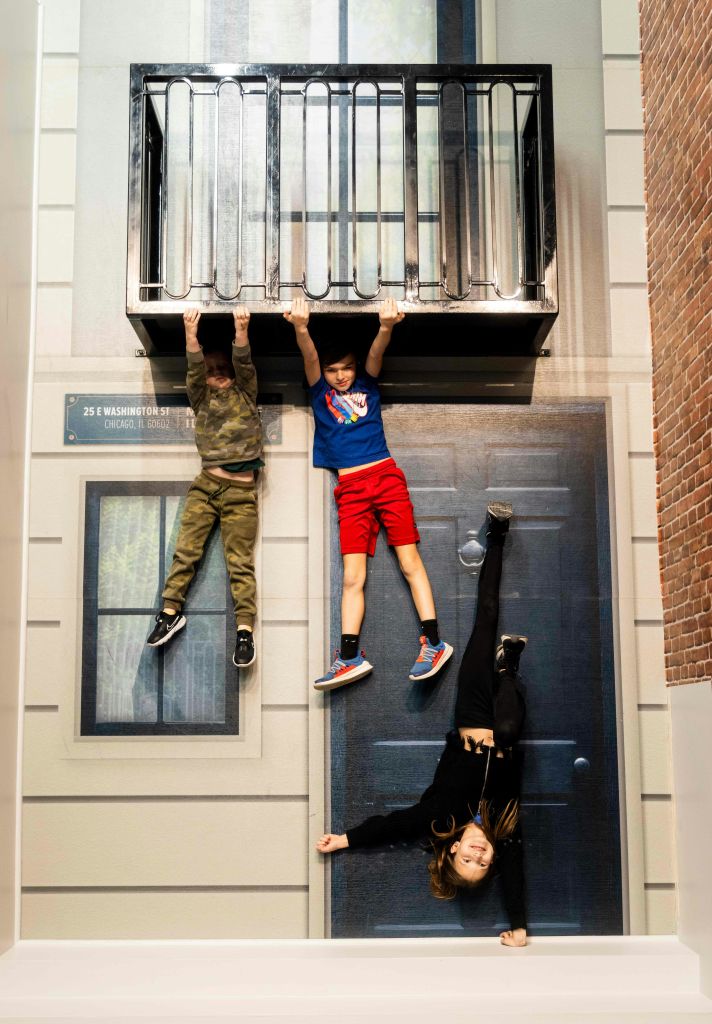 세 명의 어린이가 건물의 발코니에 매달려 있는 것처럼 보이며, 두 명은 버티고 한 명은 거꾸로 쓰러지는 척합니다.