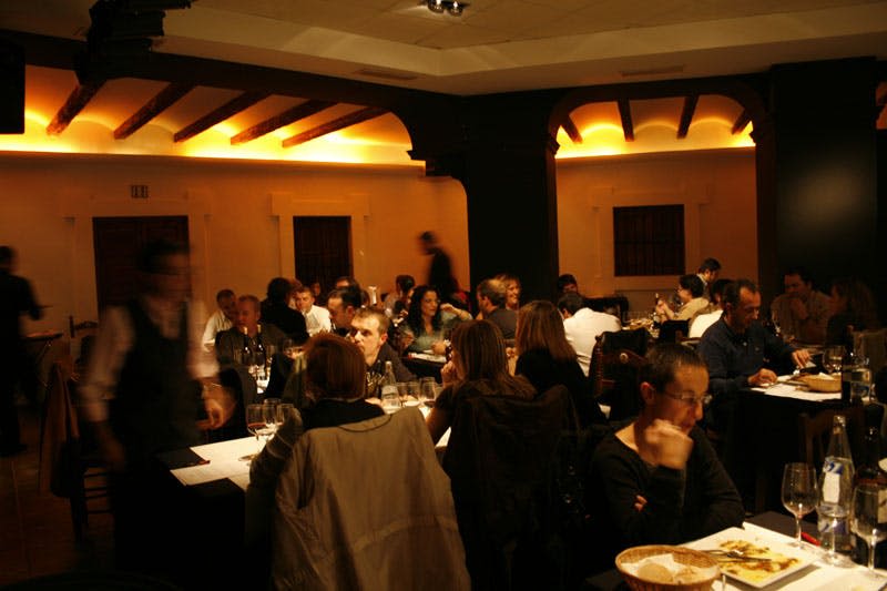 천장에 나무 기둥이 있고 앉은 손님이 많으며 웨이터가 움직이는 조명이 희미한 레스토랑입니다.