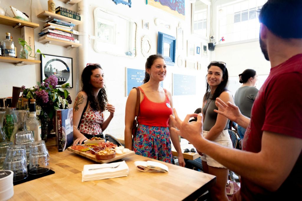 한 무리의 사람들이 카페에서 이야기를 나누고 있는데, 카운터에 음식 접시가 놓여 있고 배경에는 장식용 벽면 예술품이 놓여 있습니다.