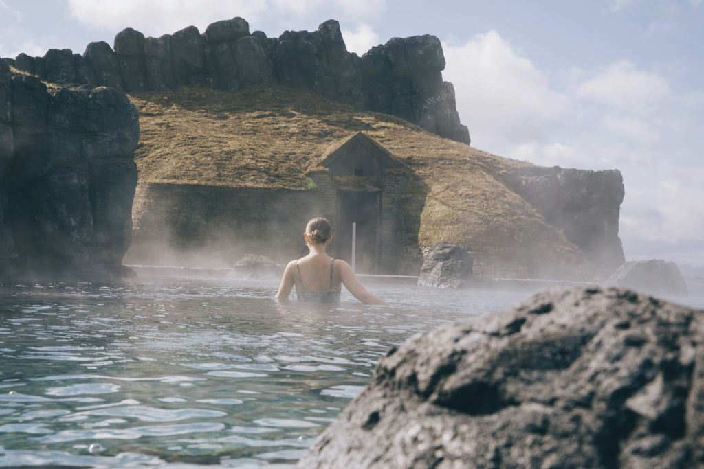 수영복을 입은 사람이 작은 석조 건물과 바위 언덕을 배경으로 안개가 자욱한 온천 수영장에 서 있습니다.