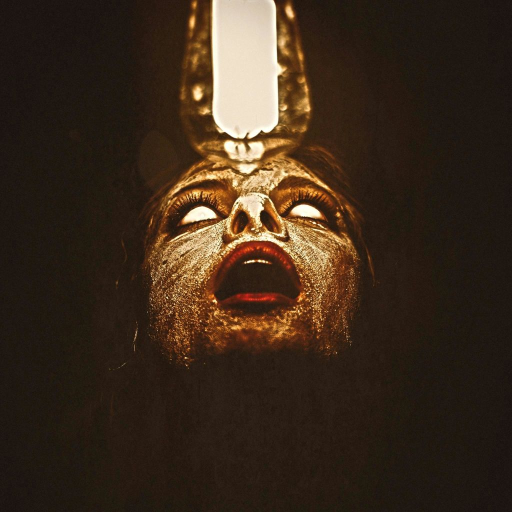 하얀 눈동자와 입을 벌린 채 위에서 비춰진 얼굴은 어두운 배경에 부족 같은 페인트로 장식되어 있습니다.