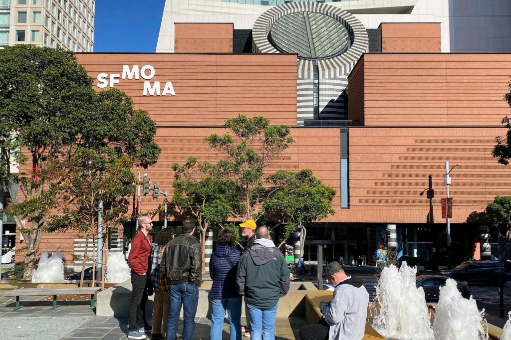 붉은 벽돌 외관, 나무, 현대적인 건축 요소가 특징인 SFMOMA 건물 밖에 서 있는 사람들의 모임.