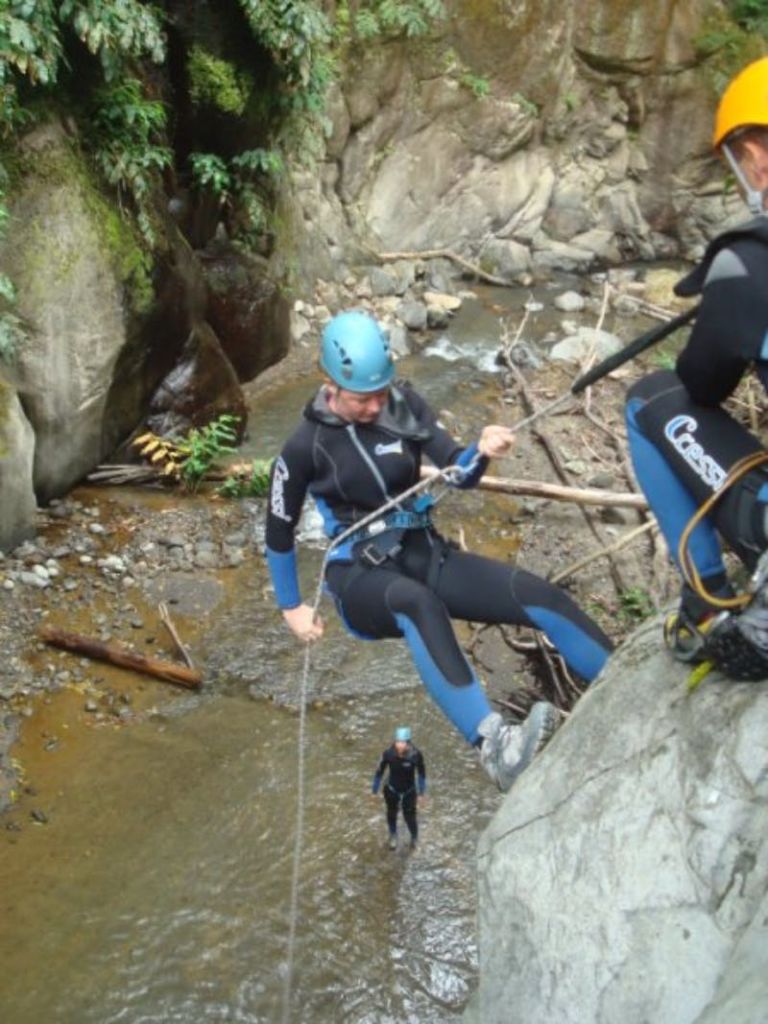 잠수복과 헬멧을 입은 사람들이 숲이 우거진 협곡의 하천 위 바위 절벽을 따라 내려오고 있습니다.