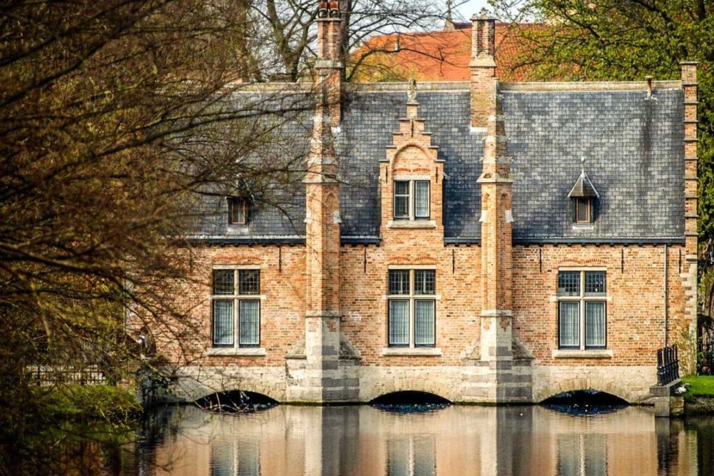 고딕 양식의 창문과 가파른 슬레이트 지붕이 있는 벽돌 건물로, 나무로 둘러싸인 눈앞의 잔잔한 물에 반사됩니다.