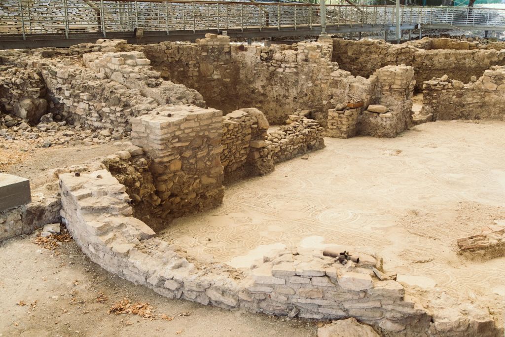 부분적으로 서 있는 벽과 눈에 띄는 모자이크 바닥이 있는 고대 석조 유적. 높은 통로를 배경으로.