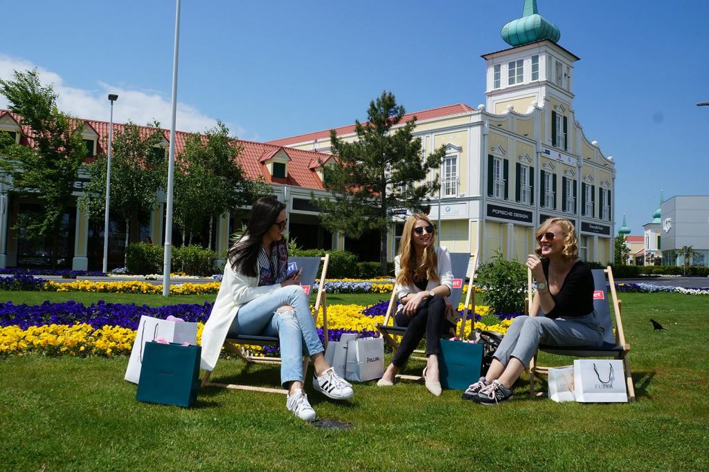 화려한 꽃과 나무가 있는 건물 밖, 쇼핑백을 들고 잔디밭에 앉아 있는 세 명의 여성.