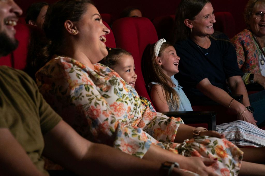 극장에 앉은 두 명의 여성과 두 명의 소녀가 빨간색 좌석과 어두운 배경으로 웃고 있습니다.