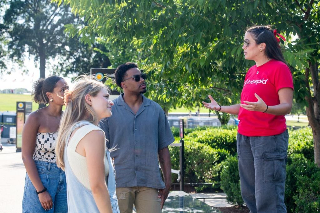야외에서 한 무리의 사람들이 햇빛 아래 녹지 근처에 서서 이야기하고 있습니다. 한 사람이 말하면서 몸짓을 하고 있습니다.