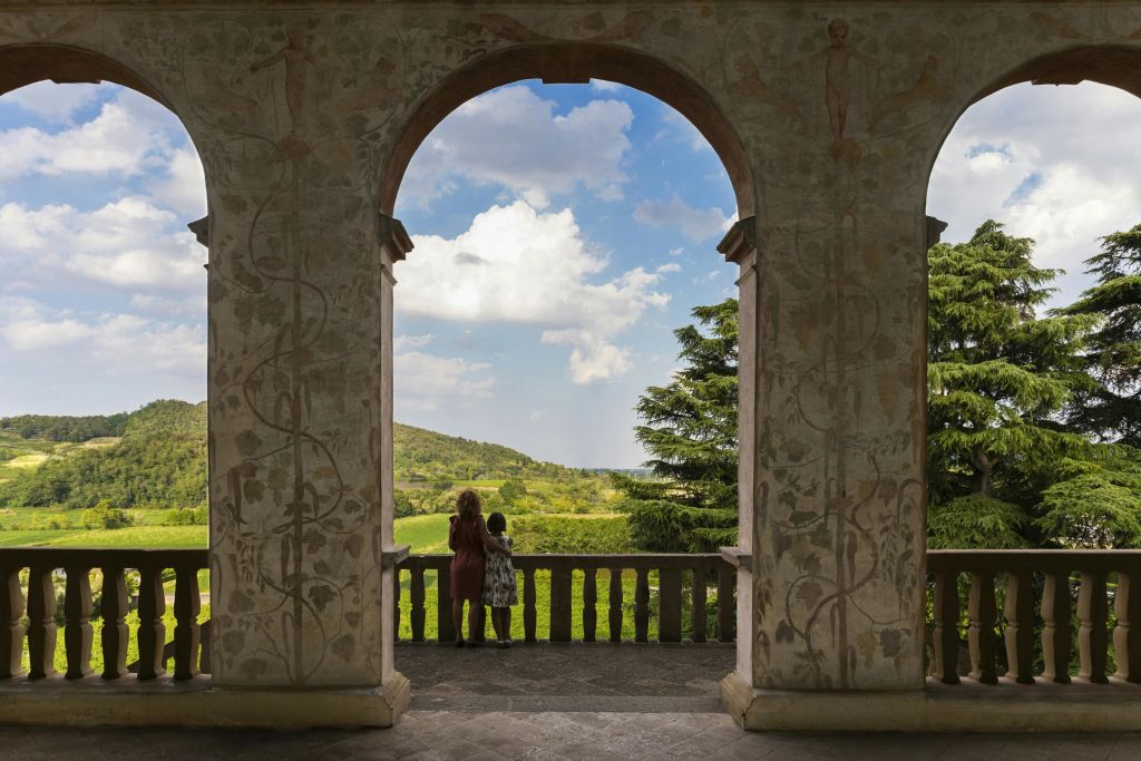 부분적으로 흐린 하늘 아래 언덕과 나무가 있는 울창하고 푸른 풍경이 내려다보이는 아치형 통로 아래 두 사람이 서 있습니다.